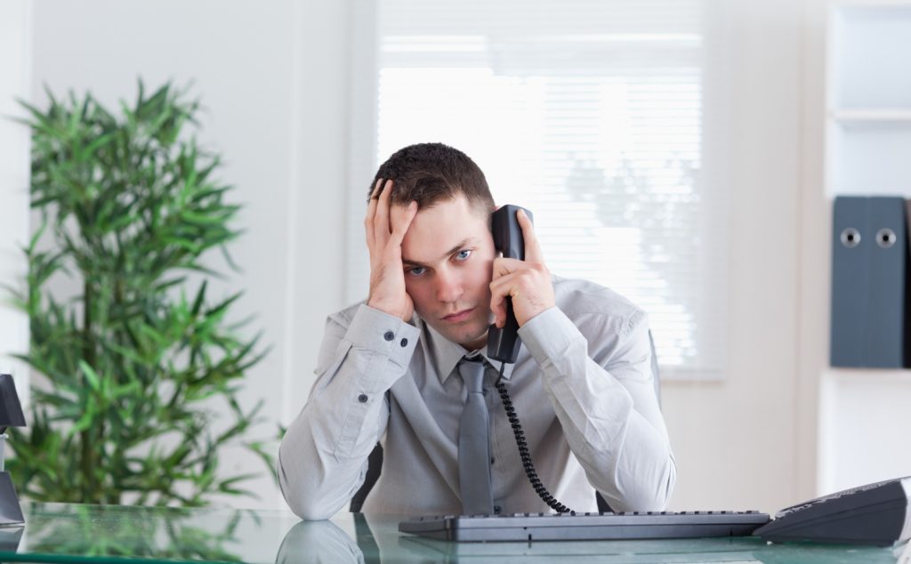 Why follow-up gets overlooked - A man in a gray shirt and tie sits at a desk, holding a phone to his ear and resting his head on one hand, appearing stressed. Office equipment and plants are visible in the background.