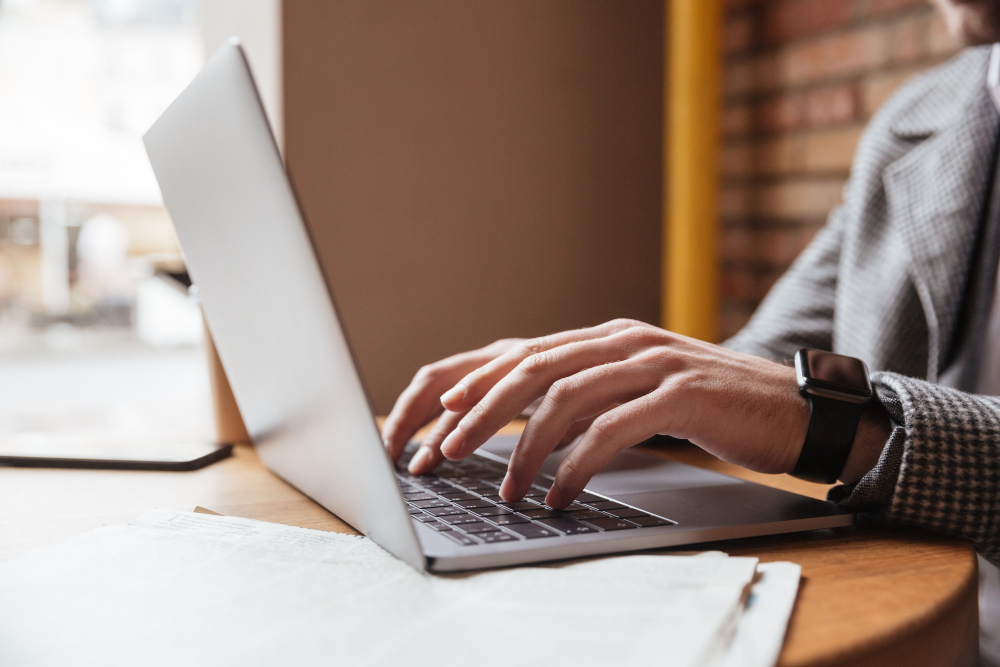 Close-up of a person in business attire typing on a laptop at a wooden table, with documents and a pen placed nearby.
