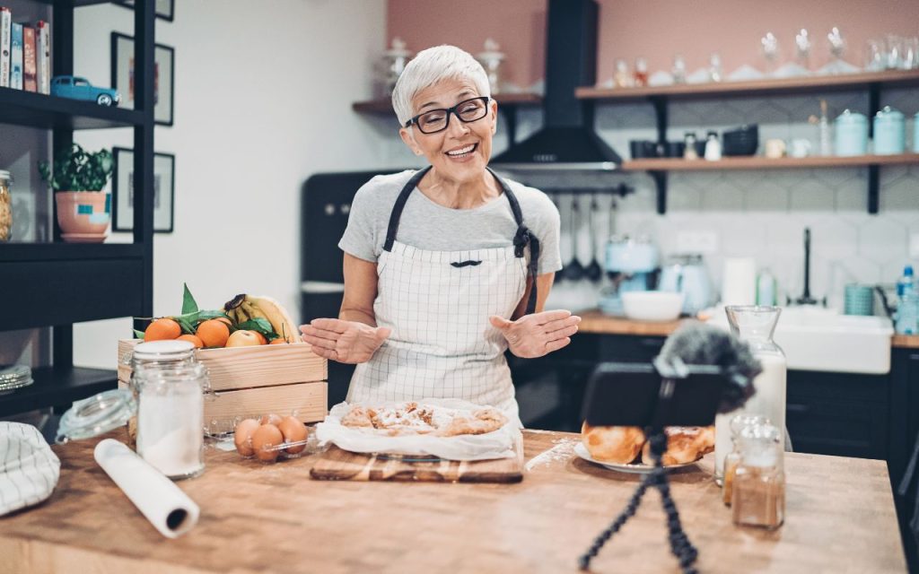 An older woman wearing glasses and an apron stands in a kitchen, smiling and gesturing to baked goods in front of a smartphone on a tripod, recording a cooking demonstration.