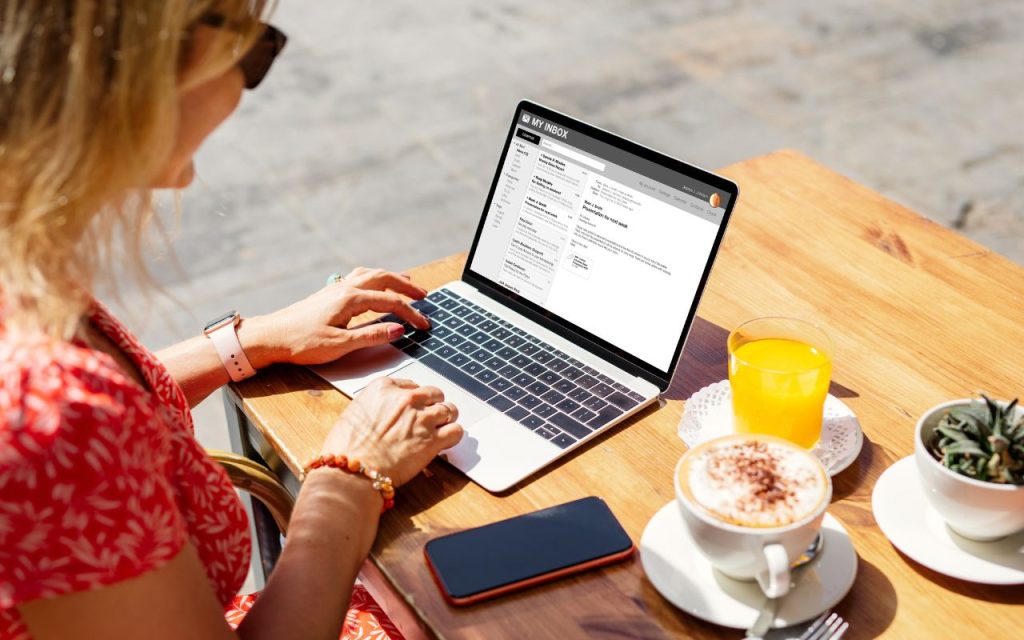 A person in a red dress uses a laptop at an outdoor table with coffee, orange juice, a smartphone, and a small potted plant. The laptop screen shows an email inbox.