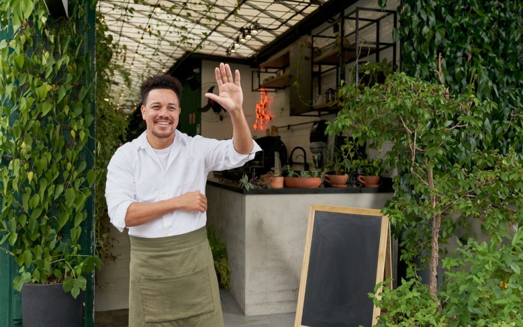 A man in a white shirt and green apron stands at the entrance of a plant-filled café, smiling and waving, with a blank chalkboard sign beside him.