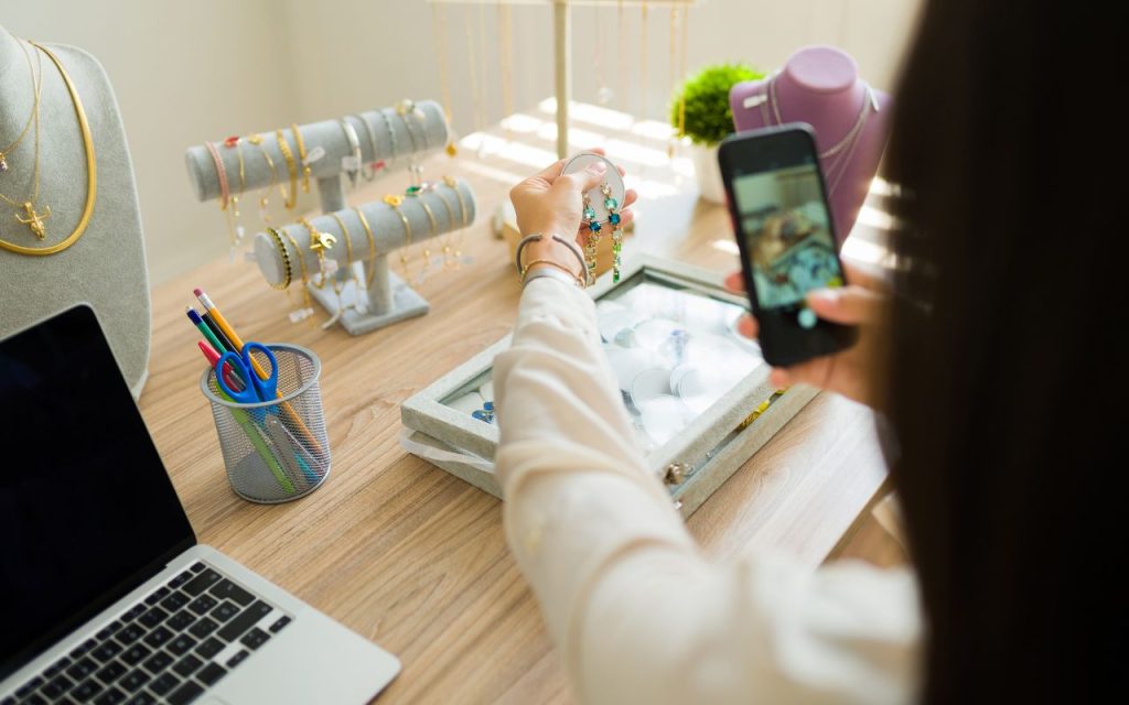 Person holds jewelry while taking a photo with a smartphone at a desk displaying assorted jewelry, a laptop, and office supplies.