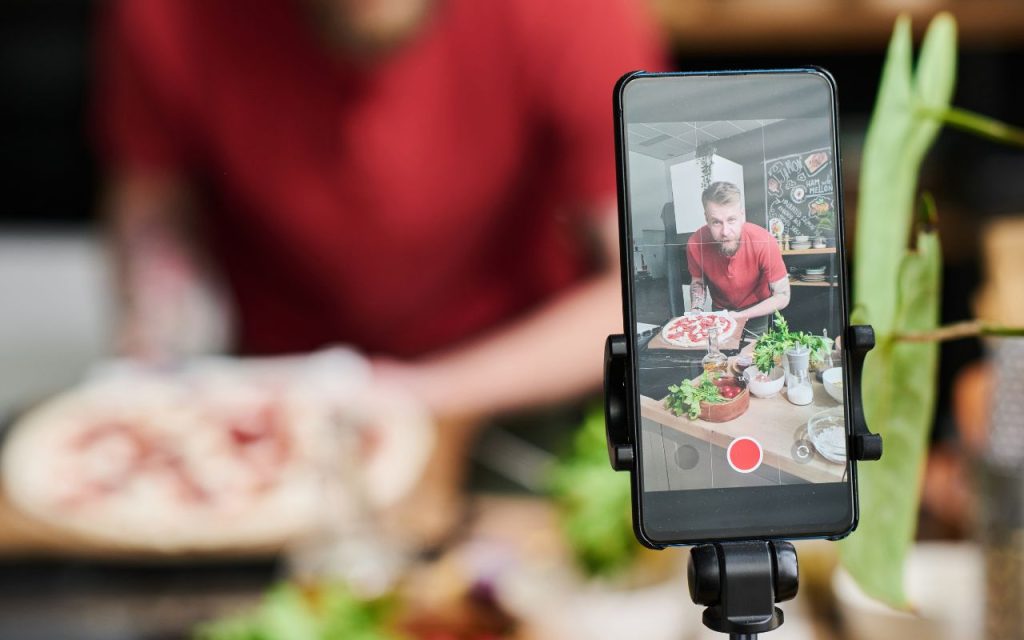 A smartphone on a tripod records a person in a red shirt preparing pizza in a kitchen with various ingredients visible on the counter.