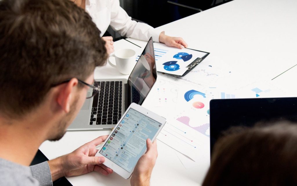Three people working at a table with laptops, a tablet, and printed charts showing business graphs and data.
