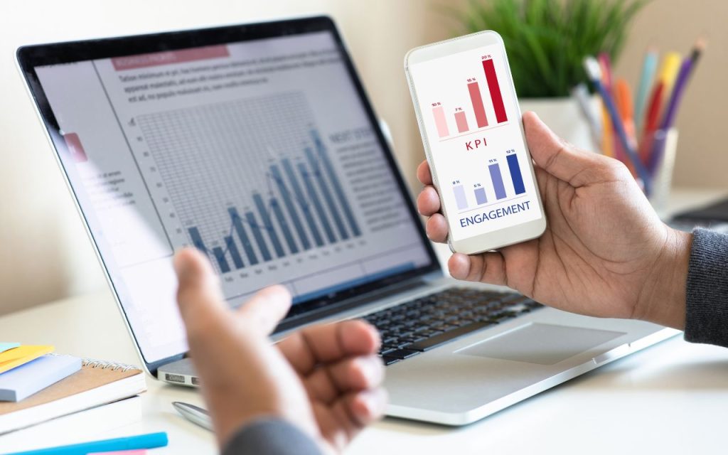 A person reviews bar graphs on a smartphone labeled "KPI" and "Engagement" while sitting at a desk with a laptop displaying a similar chart.