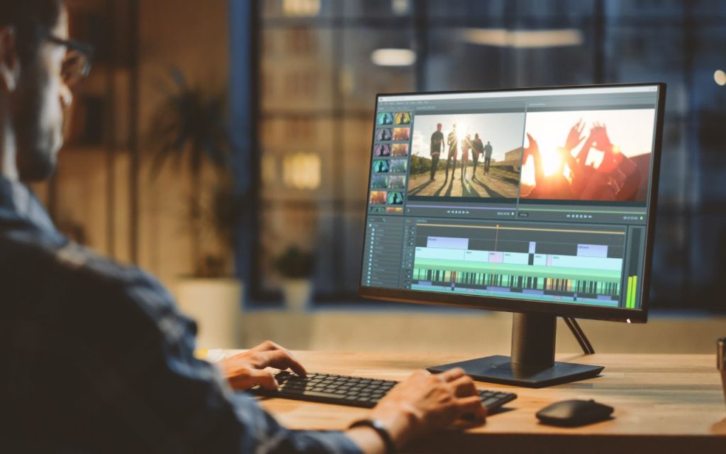 A person sits at a desk editing video footage on a computer monitor, with various clips and a timeline visible on the screen.