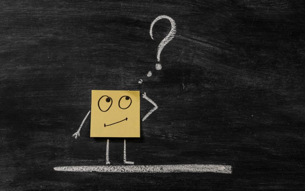 A yellow sticky note with a drawn face is placed on a blackboard, with chalk-drawn arms, legs, and a large question mark above the head.