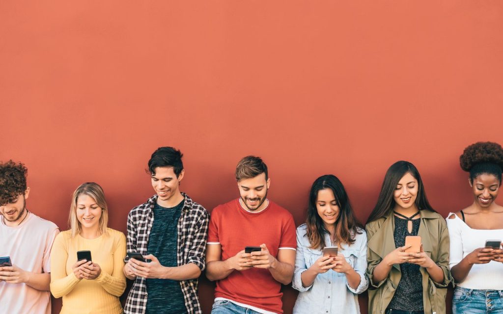 Seven young adults stand in a row against a red wall, each looking at and using their smartphone.