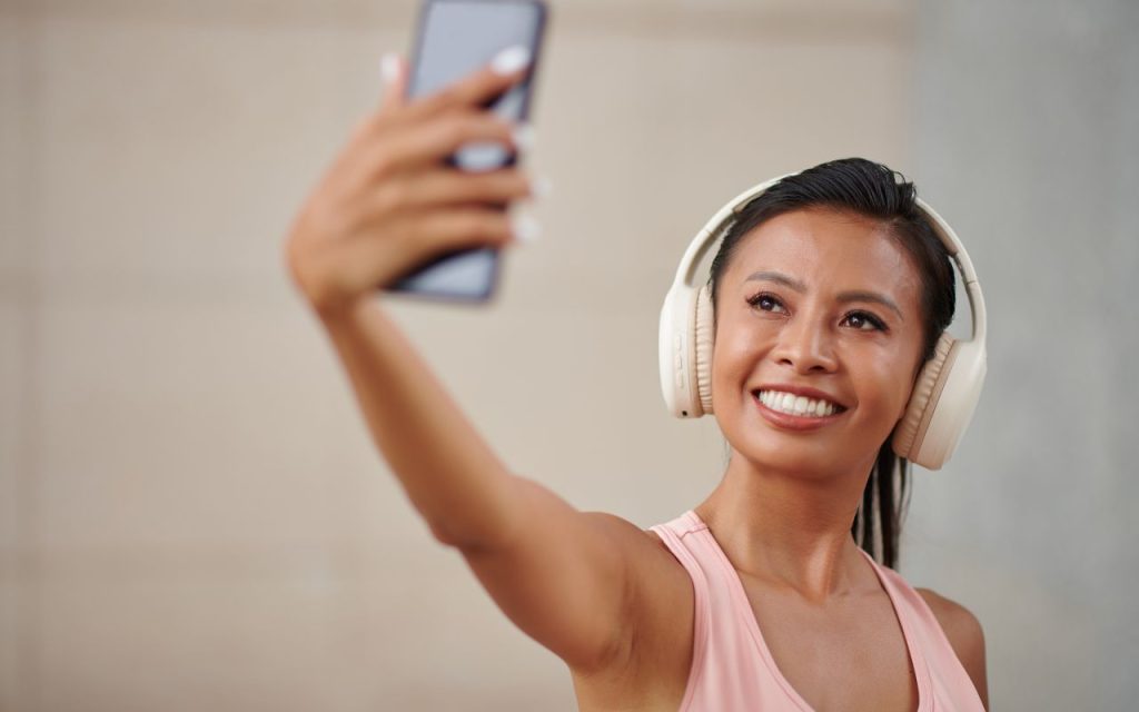 Woman wearing large headphones and a sleeveless top takes a selfie with her smartphone, smiling and looking at the camera.
