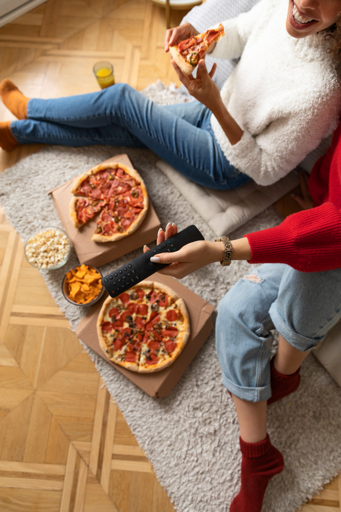 The rise of AI-powered conversational search - Two people sitting on the floor eating pizza, with open pizza boxes, snacks, and a TV remote on a rug in a casual home setting.