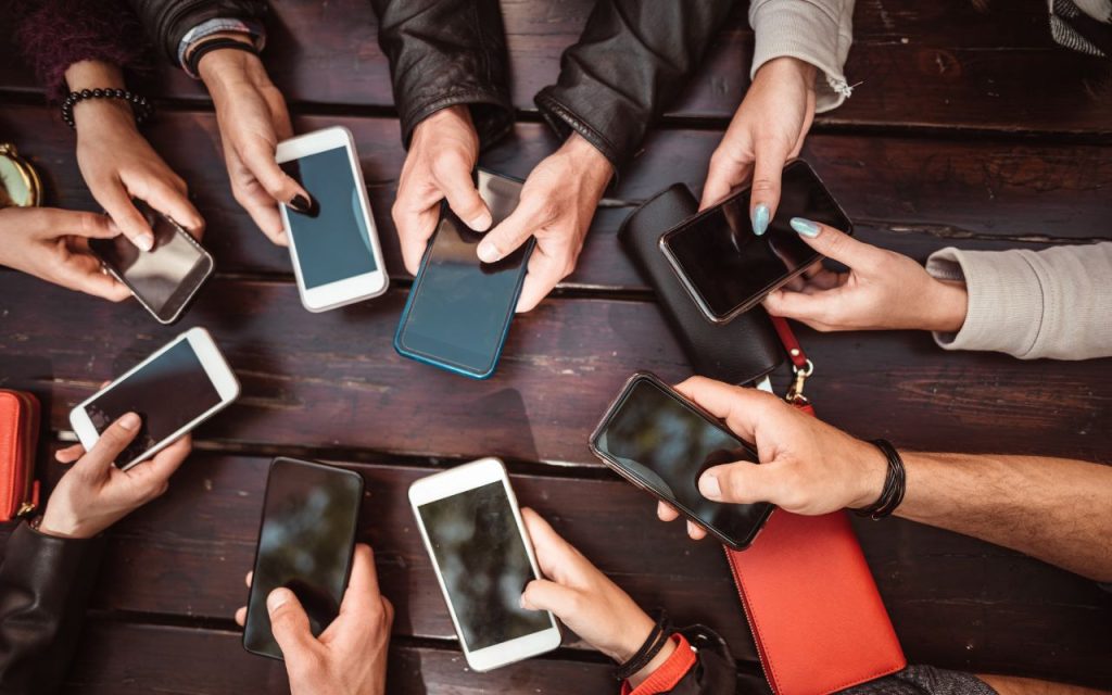 A group of people sitting around a wooden table, each holding and using a smartphone, with only their hands visible.