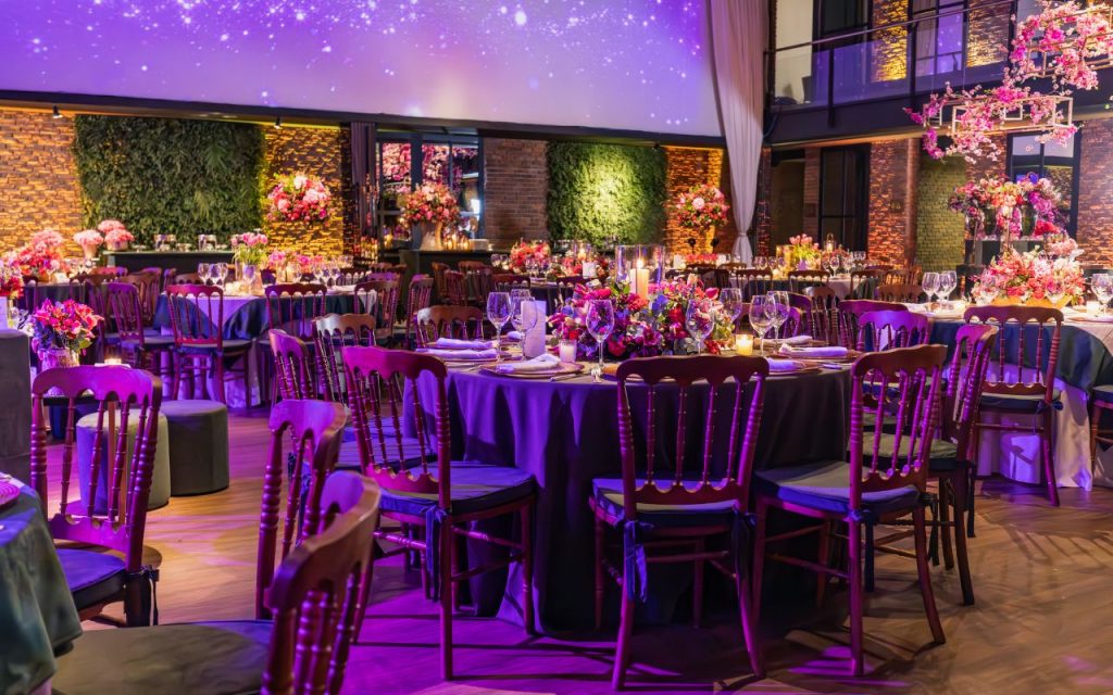 A decorated banquet hall with round tables set for an event, floral centerpieces, wooden chairs, and purple ambient lighting.