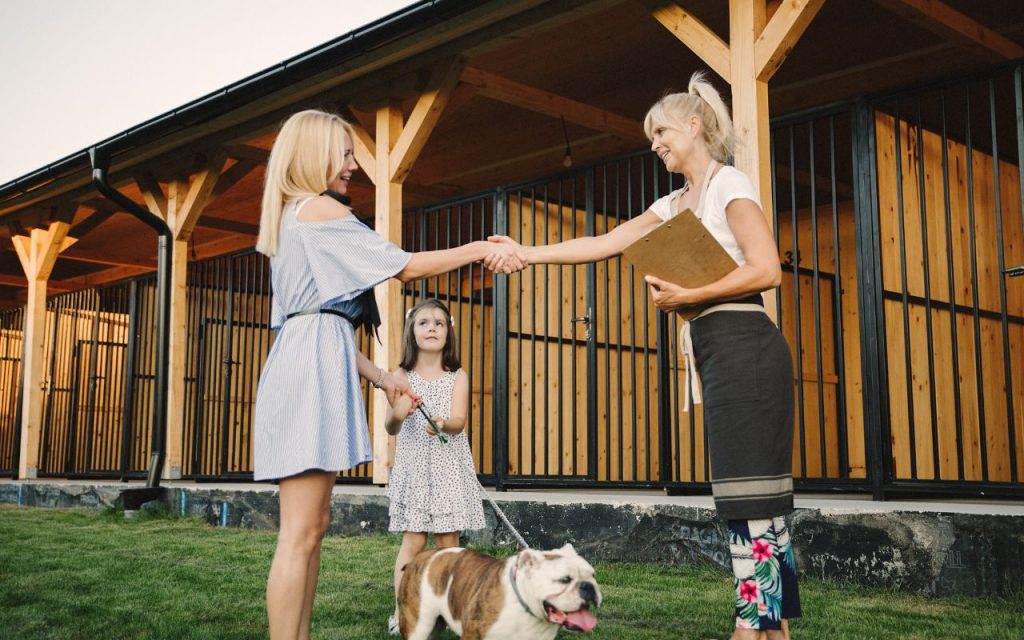 Two women shake hands outside a kennel while a young girl stands nearby holding a bulldog on a leash; one woman holds a clipboard.