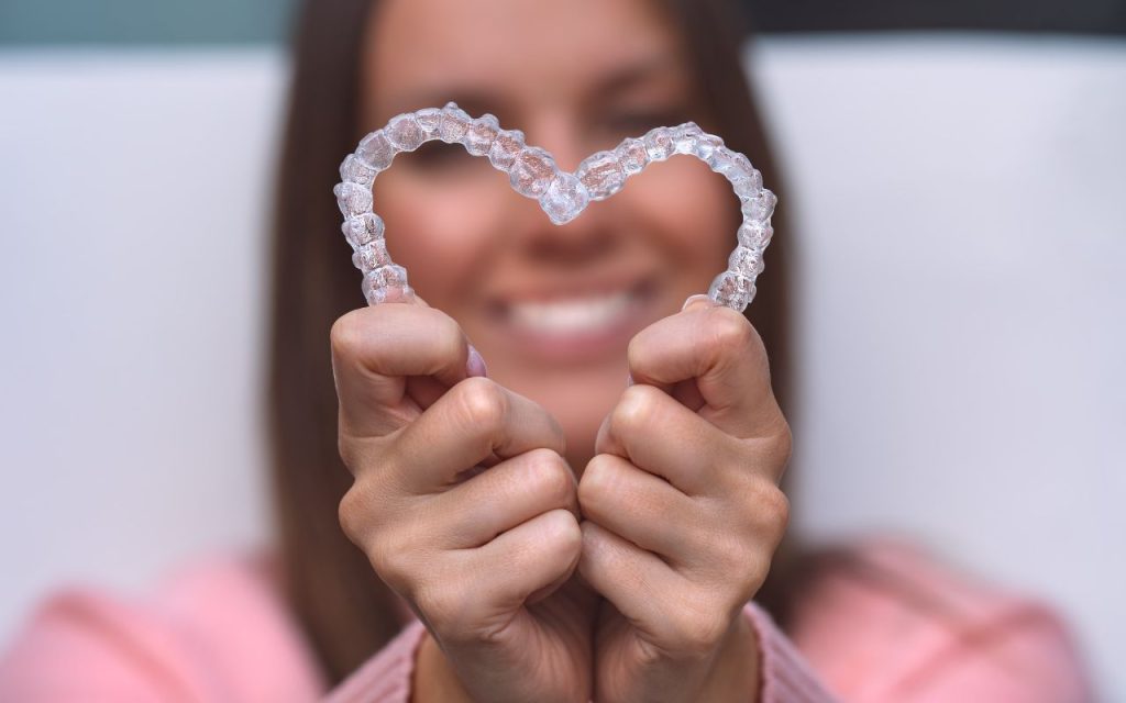 A person holds two clear dental aligners shaped into a heart, with a blurred smiling face in the background.