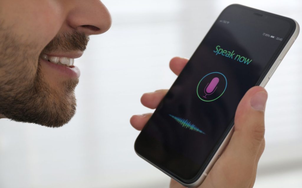 A man holds a smartphone close to his mouth, using a voice assistant app with "Speak now" and a microphone icon displayed on the screen.