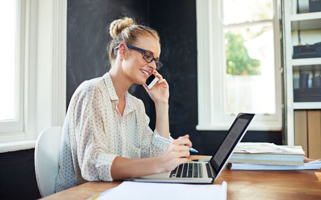 A woman wearing glasses sits at a desk, talking on a smartphone and working on a laptop, with papers and books on the table in a bright room.