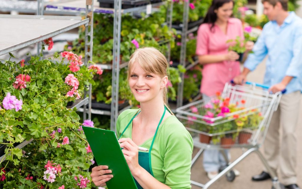 A woman in a green apron holds a clipboard and smiles in a garden center, while two people with a shopping cart select plants in the background.