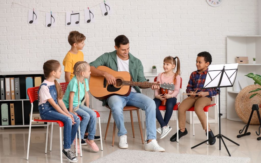 An adult teaches five children about music, playing guitar while they sit in a classroom setting with music notes on the wall and a sheet music stand nearby.