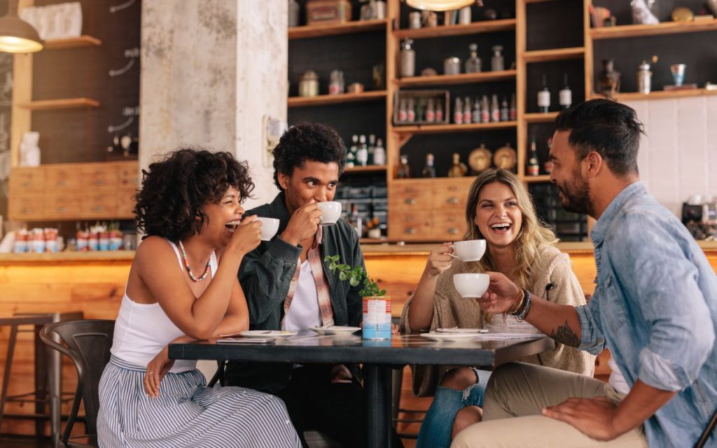 Four people sit around a table in a cafe, drinking coffee and smiling as they engage in conversation. Shelves with bottles and decor are visible in the background.