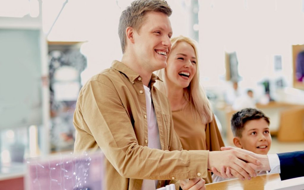 A man smiles and shakes hands across a counter, with a woman and a boy standing beside him, all appearing happy in a bright indoor setting.