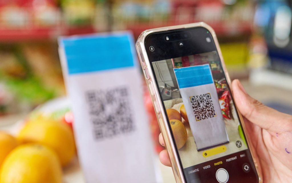 A person uses a smartphone to scan a QR code displayed on a sign at a market stall with fruit in the background.