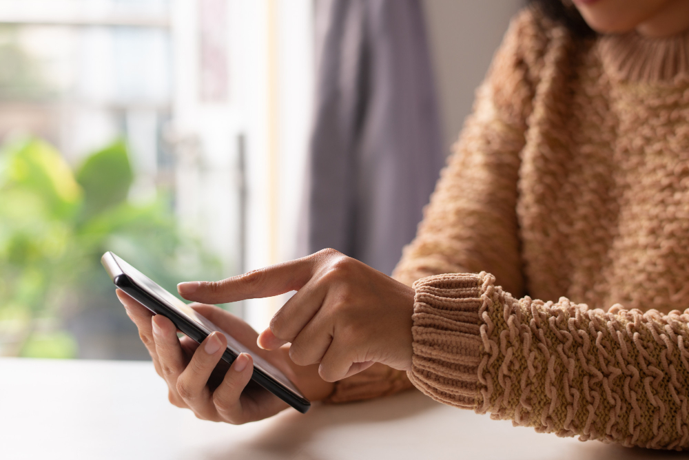 A person wearing a brown knitted sweater is using a smartphone, touching the screen with one finger while holding the device in their other hand.