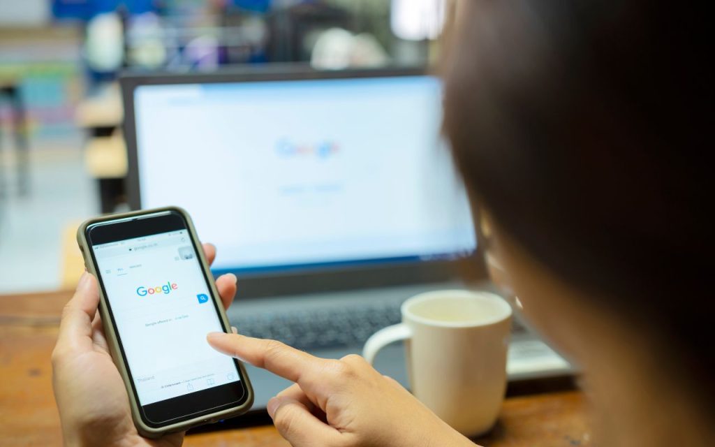 A person holds a smartphone displaying the Google homepage, with a laptop and a coffee mug in the background on a wooden table.