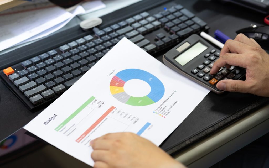 Person holding a budget report with a pie chart and using a calculator, with a computer keyboard visible on the desk.