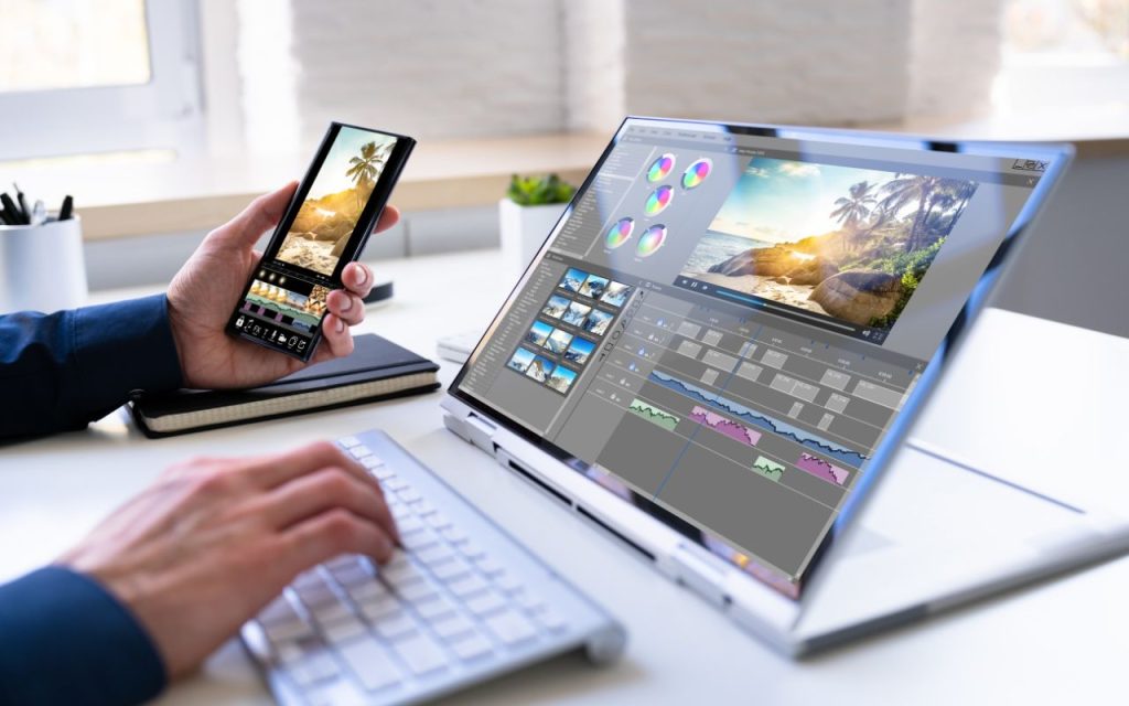 Digital marketing tools for video content. A person edits a tropical beach photo on a laptop using photo editing software, while holding a smartphone displaying the same image.