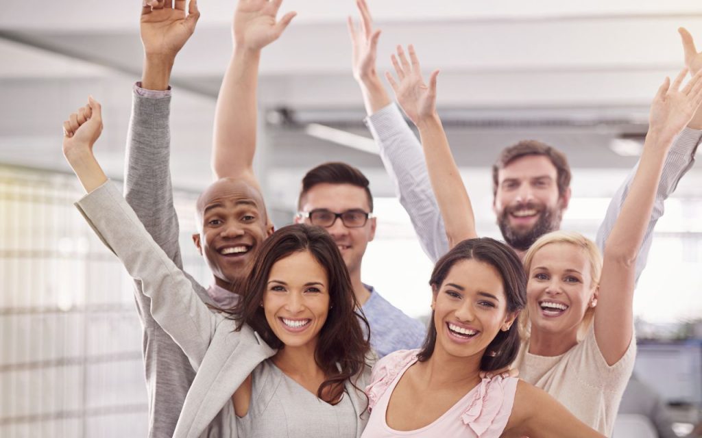 Six people stand together indoors, smiling and raising their arms in celebration or excitement.