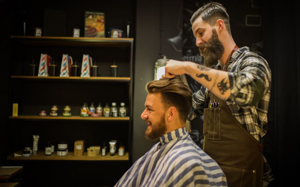 social-media-strategy-show-the-work-videos - A barber styles a smiling customer's hair in a barbershop; hair products are visible on shelves in the background.