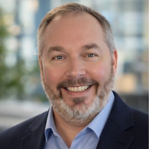 Smiling man with a beard and short hair wearing a dark suit jacket and light blue shirt, standing in front of a blurred indoor background.