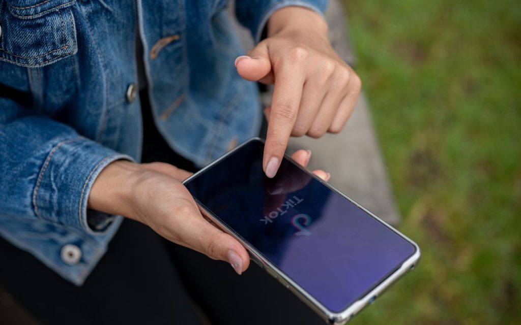 A person holds a smartphone and uses their finger to open the TikTok app; they are wearing a denim jacket and sitting outdoors.