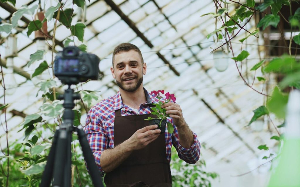 What to actually film for TikTok Local Feed - A man in an apron holds a potted plant and smiles at a camera on a tripod in a greenhouse filled with leafy plants.